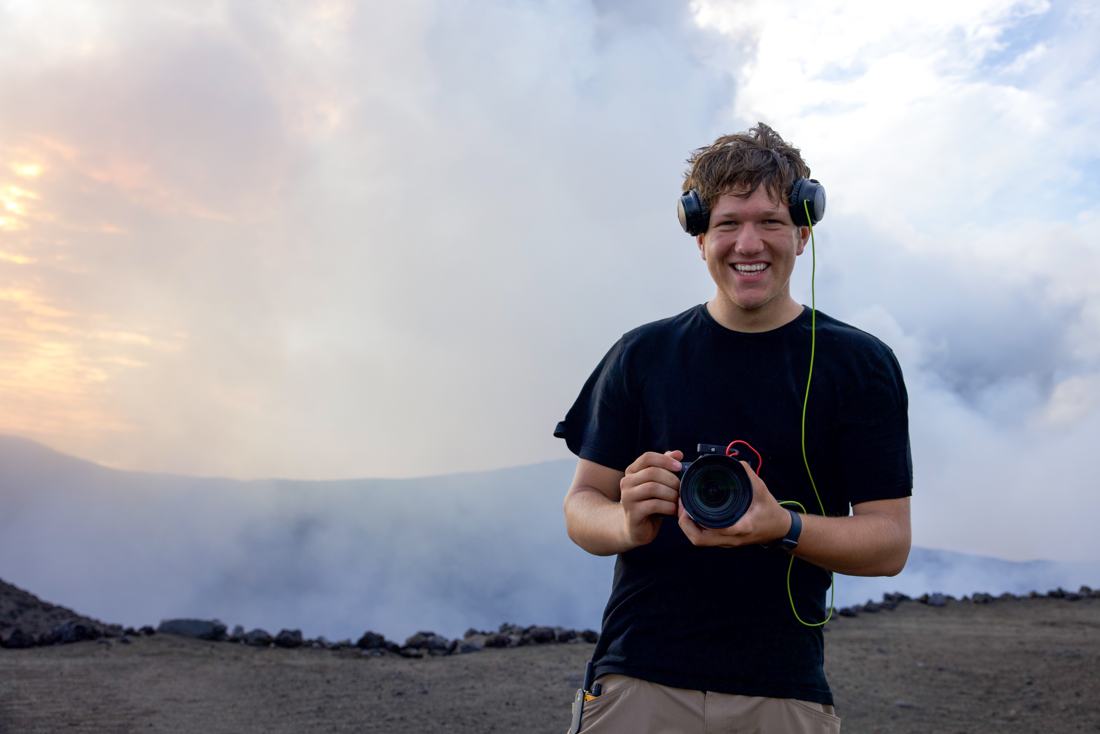 Gabriel smiling with a camera and headphones at the rim of a smoking volcano crater